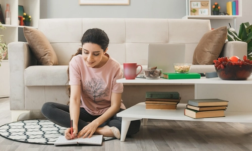 Pleased young girl writes on notebook sitting on floor behind coffee table in living room