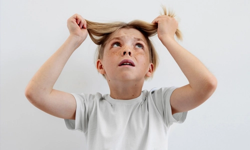 A young child looks upward with a distressed expression while pulling at their hair with both hands.
