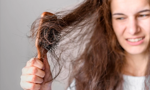 A woman with a distressed facial expression holds a wooden hairbrush tangled in her messy, frizzy brown hair.